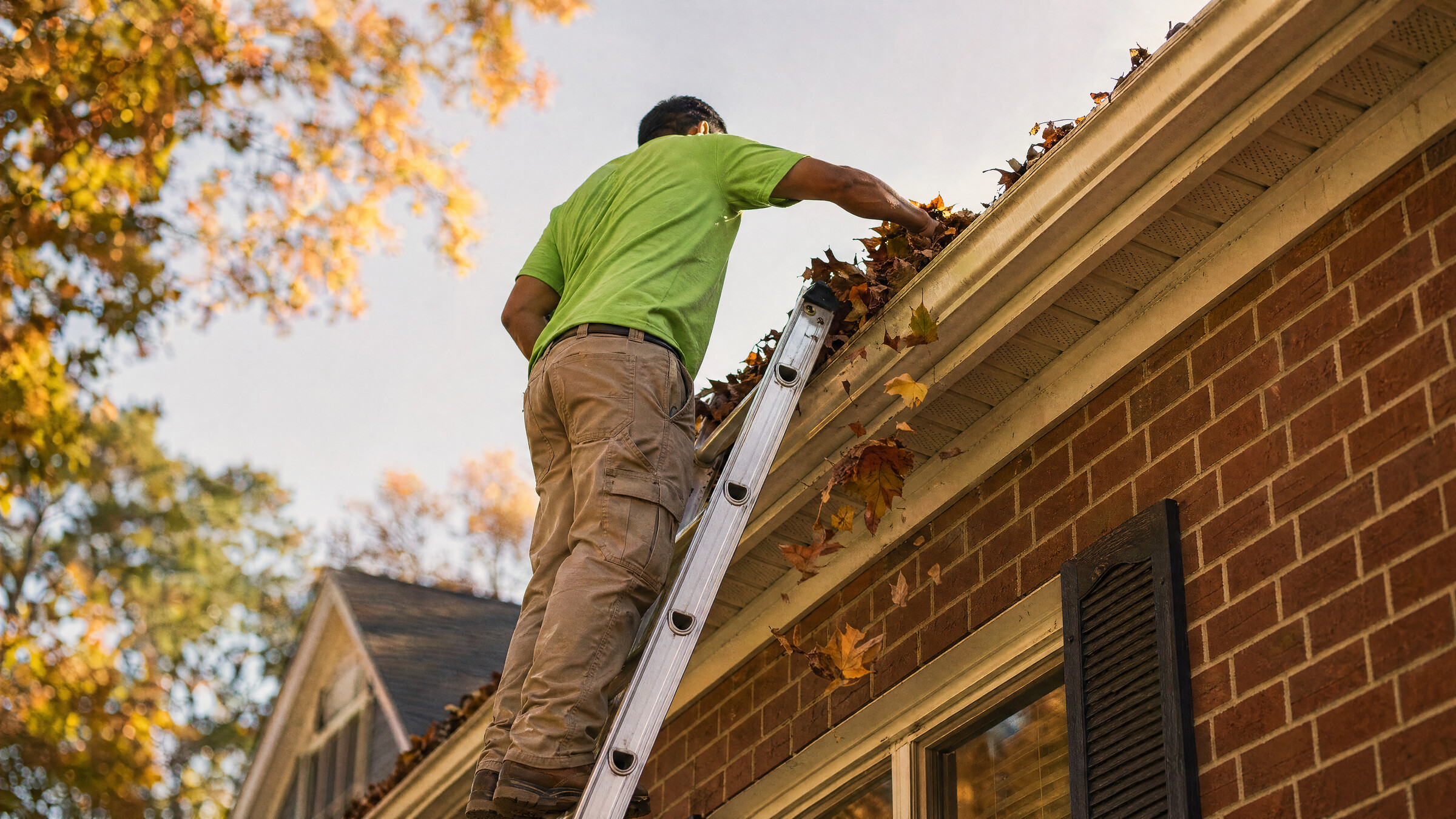 Contractor on an extension ladder clearing gutters on a two-story brick Durham home